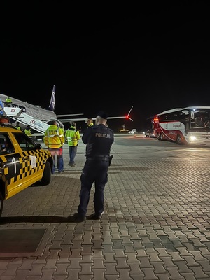 policjant w niebieskim mundurze, w tle samolot. Z prawej strony autokar z napisem Łączy nas piłka