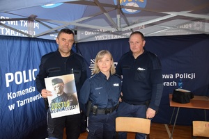 dwóch policjantów i policjantka wewnątrz niebieskiego namiotu, policjant trzyma plakat z napisem &lt;zostań policjantem&gt;