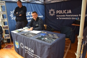 policjant siedzący przy stole znajdującym się pod namiotem policyjnym, obok niego inny policjant