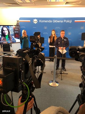 Policjant na tle niebieskiej ścianki z napisem Komenda Główna Policji, obok inne osoby, telewizor, kamery