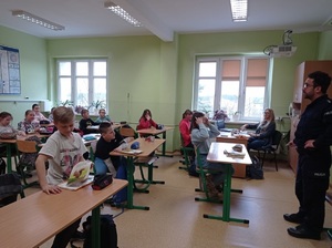 pomieszczenie szkolnej klasy, w której uczniowie siedzący w ławkach słuchają prelekcji policjanta