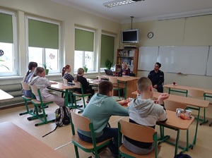 pomieszczenie szkolnej klasy, w której uczniowie siedzący w ławkach słuchają prelekcji policjanta