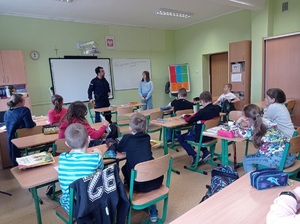 pomieszczenie szkolnej klasy, w której uczniowie siedzący w ławkach słuchają prelekcji policjanta