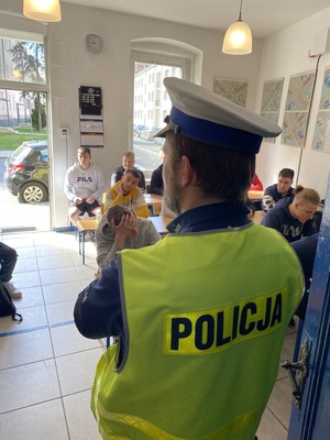 policjant w odblaskowej kamizelce i białej czapce, przed nim uczestnicy konkursu siedzący w ławkach