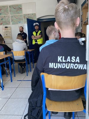 na pierwszym planie uczestnik konkursu, siedzący na krześle, w czarnej koszulce z napisem klasa mundurowa, w tle inni uczestnicy i policjanci w mundurach