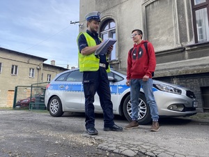 policjant w niebieskim mundurze, odblaskowej kamizelce i białej czapce, obok niego uczestnik konkursu w czerwonej bluzie, za nimi policyjny radiowóz