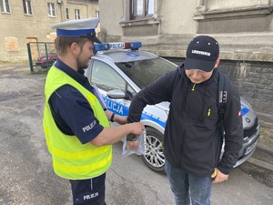 policjant trzyma w rękach woreczek z pytaniami konkursowymi, pytanie losuje uczestnik w ciemnym ubraniu, w tle radiowóz