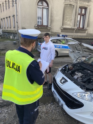 policjant i uczestnik konkursu przy białym pojeździe z otwartą pokrywą silnika, w tle radiowóz i inny czarny pojazd