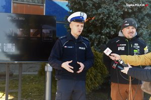 policjant w mundurze w trakcie wypowiedzi, obok telewizor i inny mężczyzna w ubiorze cywlinym