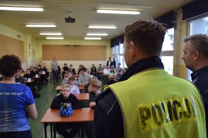 policjant w kamizelce odblaskowej, w tle uczniowie w ławkach