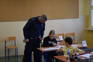 policjant w niebieskim mundurze pochylający się nad stolikiem przy którym siedzi uczestnik konkursu