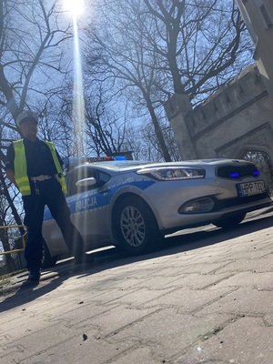 policyjny radiowóz przy którym stoi policjant w niebieskim mundurze, białej czapce i kamizelce odblaskowej