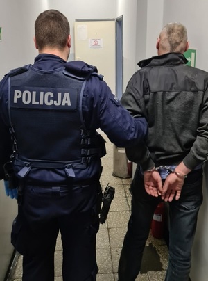 policjant w niebieskim mundurze prowadzący korytarzem mężczyznę skutego kajdankami na ręce z tyłu