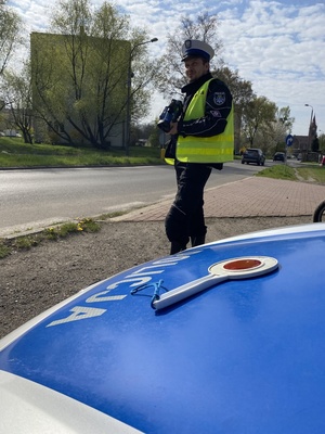 maska policyjnego radiowozu koloru niebieskiego, na której leży tarcza biało czerwona, do zatrzymywania pojazdów, w tle policjant