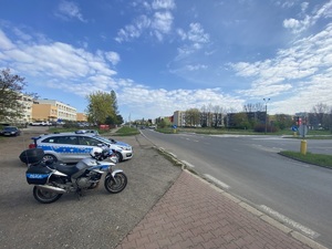 skrzyżowanie ulic, przy jezdni policyjny radiowóz i motocykl, w tle zabudowania i pojazdy
