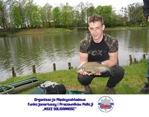 mężczyzna trzymający w rękach rybę, w tle zbiornik wodny, w dolnej części logo organizacji związkowej