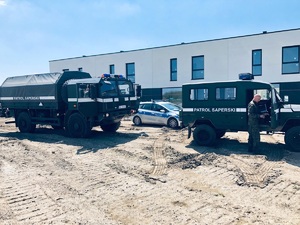 radiowóz policyjny oraz dwa pojazdy wojskowe z napisem patrol saperski, w tle budynki