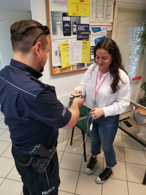 policjant w mundurze wręczający kobiecie dokumenty w pomieszczeniu, za nimi tablica informacyjna