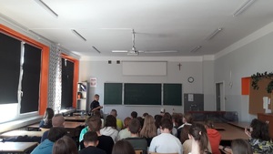 uczniowie w szkolnych ławkach, przed nimi policjantka w mundurze