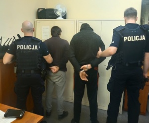 w pomieszczeniu dwóch policjantów trzymających za ręce dwóch zatrzymanych skutymi kajdankami, w tle szafa
