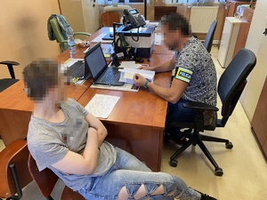 policjant w ubiorze cywilnym w opaska odblaskową z napisem policja siedzący przy biurku. Obok siedzącą kobieta w jasnym ubiorze