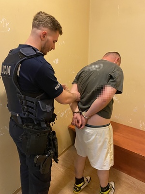 policjant trzymający skutego kajdankami zatrzymanego w pomieszczeniu komendy
