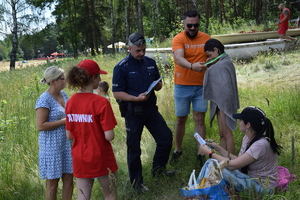 policjant, ratownik i przedstawiciele gminy Świerklaniec rozmawiający z osobami wypoczywającymi na trawie