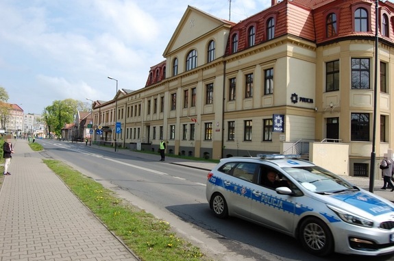 Fotografia kolorowa. Na zdjęciu widać budynek Komendy Powiatowej Policji w Tarnowskich Górach, obok którego przejeżdża policyjny radiowóz.