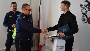 tarnogórski komendant wręczający podziękowania i gratulujący panu Łukaszowi.  W tle umundurowany zastępca naczelnika wydziąłu ruchu drogowego