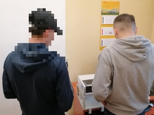 mężczyzna ubrany na czarno z zamazaną twarzą, obok stojący policjant po cywilnemu wykonujący czynności