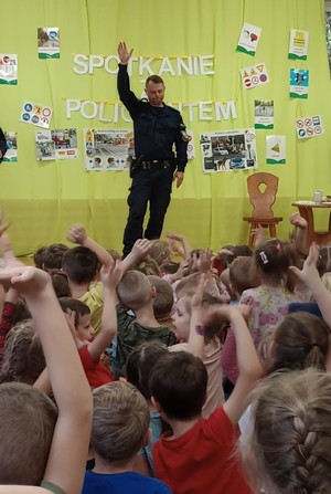 Policjant podczas spotkania z przedszkolakami. policjant z podniesioną ręką. Dzieci odwróceni tyłem do obiektywu.
