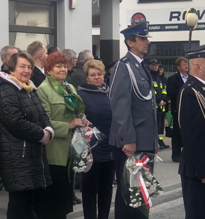 policjant w umundurowaniu galowym, w ręku trzyma wiązankę kwiatów, obok postronne osoby