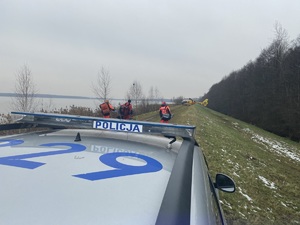 na zdjęciu dach policyjnego radiowozu, za którym idą ratownicy w stronę stojącego dalej helikoptera ratowniczego. Wszystko przy zbiorniku wodnym