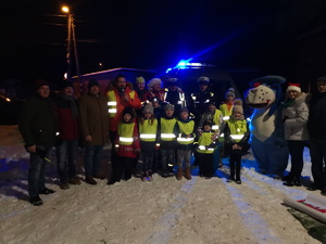na zdjęciu dzieci w odblaskowych kamizelkach, obok maskotka policyjna, dorośli w tle radiowóz