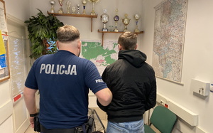 na zdjęciu policjant w mundurze, w pomieszczeniu stoi i trzyma zatrzymanego za ramię