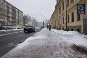 na zdjęciu zaśnieżony chodnik, na nim pieszy, w około budynki i samochody