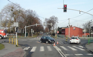 na zdjęciu skrzyżowanie ulic z sygnalizacją świetlną, na którym stoi rozbity czarny samochód, a obok strażak kieruje ruchem