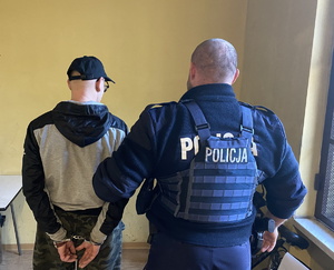 na zdjęciu policjant w pomieszczeniu komendy, który trzyma za rękę zatrzymanego, który na rękach z tyłu ma kajdanki