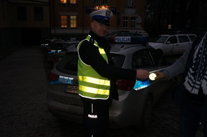 na zdjęciu policjant wręcza osobie odblask, za nim samochody i budynki