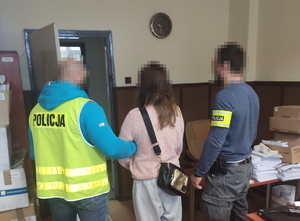 na zdjęciu policjanci w kamizelce odblaskowej i odblaskowej opasce w pomieszczeniu, pomiędzy nimi zatrzymana kobieta