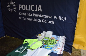 na zdjęciu fragment namiotu informacyjnego z napisem komenda powiatowa policji w tarnowskich górach, na pierwszym planie stolik z gadżetami