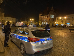 na zdjęciu policyjny radiowóz, obok którego stoją policjanci, w tle, na płycie rynku, inne osoby