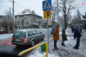 na zdjęciu fragment jezdni, po której jedzie pojazd, obok na chodniku policjant rozmawia z dziećmi i ich rodzicami