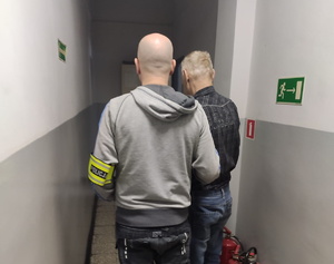 na zdjęciu policjant z opaską na ramieniu z napisem &quot;policja&quot;, prowadzi korytarzem zatrzymanego