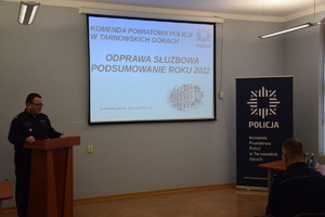 na zdjęciu komendant powiatowy przy mównicy w sali narad, w centralnej części fotografii na ekranie wyświetlana prezentacja multimedialna