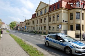 na zdjęciu policyjny radiowóz na jezdni, za nim budynek komendy, przy którym na chodniku stoi policjant, w oddali inne zabudowania