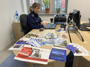 na zdjęciu policjantka przy biurku, obok niej ulotki informacyjne