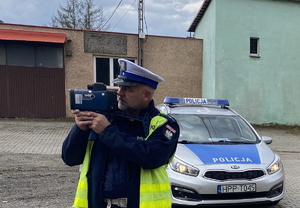 na zdjęciu policjant stojący przed zaparkowanym radiowozem i prowadzi pomiar prędkości, w rękach trzymając fotoradar