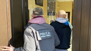 na zdjęciu policjant w ubiorze cywilnym z napisem na plecach &#039;Policja Wydział Kryminalny&quot; prowadzi zatrzymanego przez główne wejście do budynku komendy