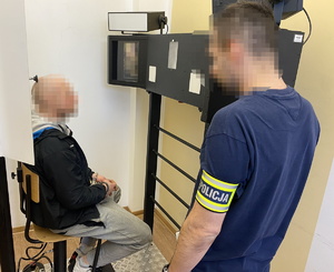 na zdjęciu technik kryminalistyki z opaską na ramieniu z napisem &#039;policja&#039;, przed którym siedzi zatrzymany w trakcie